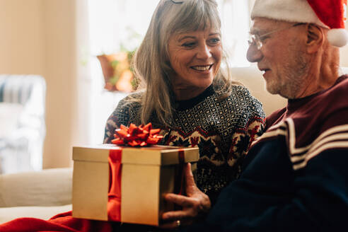 Ältere Frau schenkt ihrem Mann ein Weihnachtsgeschenk. Älteres Paar tauscht Geschenke aus und feiert Weihnachten zu Hause. - JLPSF23228