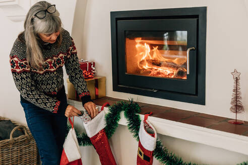 Frau, die Sachen in Weihnachtssocken steckt, während sie ihr Haus für Weihnachten dekoriert. Ältere Frau, die ihr Haus für Weihnachten dekoriert, steht vor einer Feuerkammer. - JLPSF23229