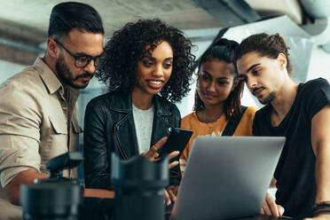 Fotograf mit seinem Team, der sich die Fotos auf einem Laptop ansieht. Model mit Handy in der Hand, das mit den Fotografen steht und sich die Fotos ansieht. - JLPSF23261