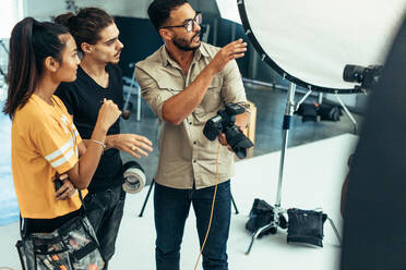 Fotograf, der seinem Team im Studio die Aufnahme erklärt. Fotograf, der mit seinen Assistenten spricht, die während eines Fotoshootings eine Kamera halten. - JLPSF23274