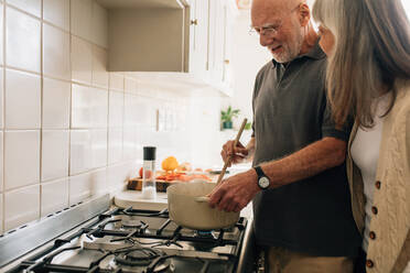 Älterer Mann, der Essen kocht, während seine Frau mit ihm spricht. Älteres Paar, das Zeit miteinander verbringt und Essen kocht. - JLPSF23276