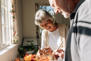 Fröhliche alte Frau, die Gemüse schneidet, während ihr Mann das Essen kocht. Älteres Paar, das Spaß daran hat, gemeinsam zu Hause zu kochen. - JLPSF23277