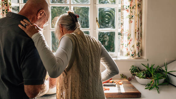 Rear view of a senior couple standing together in kitchen cooking food. Elderly woman watching her husband cook food standing in kitchen. - JLPSF23278