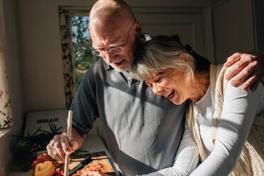 Älterer Mann, der Essen kocht und seine Frau im Arm hält, in der Küche stehend. Älteres Paar, das eine gute Zeit hat, indem es zusammen zu Hause Essen kocht. - JLPSF23280