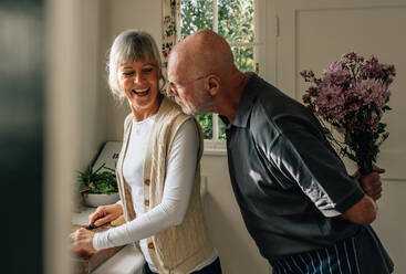 Älterer Mann versteckt einen Blumenstrauß hinter sich, um seine Frau zu überraschen. Frau schneidet Gemüse in der Küche, während ihr Mann mit einem Blumenstrauß hinter ihr steht. - JLPSF23285