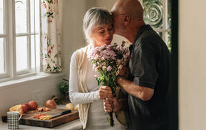 Älteres Ehepaar steht in der Küche und hält einen Blumenstrauß. Älterer Mann küsst seine Frau und hält ihre Hand zu Hause. - JLPSF23286