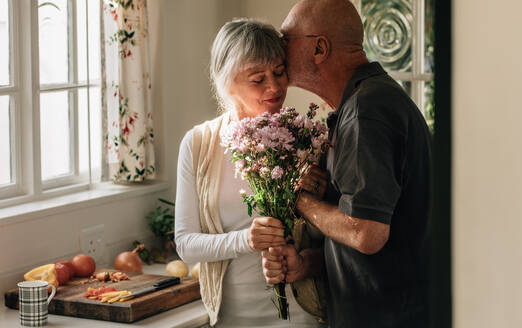 Älteres Ehepaar steht in der Küche und hält einen Blumenstrauß. Älterer Mann küsst seine Frau und hält ihre Hand zu Hause. - JLPSF23286