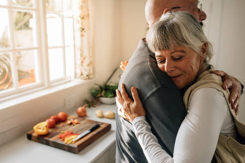 Seitenansicht eines älteren Paares, das sich zu Hause umarmt. Ältere Frau umarmt ihren Mann mit geschlossenen Augen in der Küche stehend. - JLPSF23290