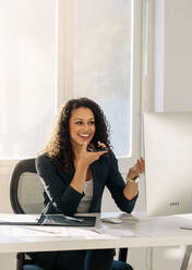 Woman sitting in front of computer in office discussing business on cell phone. Smiling woman entrepreneur at her desk in office talking over mobile phone on speaker. - JLPSF23295