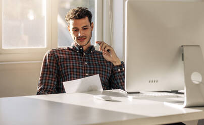 Mann, der an seinem Schreibtisch im Büro sitzt und ein Geschäftspapier betrachtet. Unternehmer, der vor einem Computer im Büro sitzt und ein Papier liest. - JLPSF23301