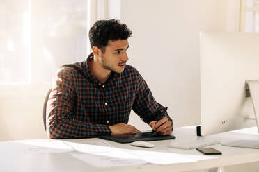 Mann sitzt an seinem Schreibtisch im Büro und schreibt auf einem digitalen Schreibblock und schaut auf den Computer. Unternehmer macht Notizen mit elektronischem Schreibblock vor einem Computer im Büro sitzend. - JLPSF23302