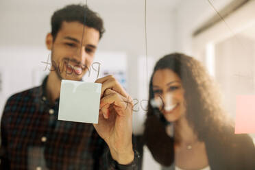 Ein Ehepaar schreibt auf Haftnotizen, die auf eine transparente Glaswand im Büro geklebt sind. Bürokollegen diskutieren Geschäftsideen und Pläne auf einer transparenten Glastafel. - JLPSF23312