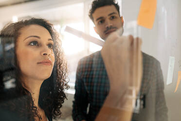 Office colleagues discussing business ideas and plans on a transparent glass wall. Business couple writing on sticky notes pasted on glass board in office. - JLPSF23314