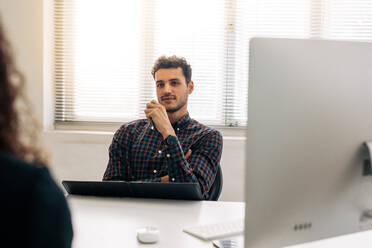 Businessman discussing work with a business partner sitting at his desk in office. Business partners in a meeting in office. - JLPSF23327