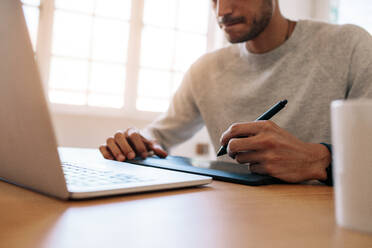 Mann schreibt auf einem digitalen Schreibblock, der an einen Laptop angeschlossen ist, der auf einem Tisch im Büro sitzt. Mann schreibt auf einem elektronischen Schreibblock mit digitalem Stift. - JLPSF23334