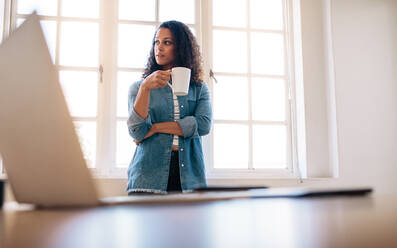 Eine Unternehmerin steht in einem Büro und hält eine Kaffeetasse, während sie wegschaut. Eine Frau steht in der Nähe ihres Tisches und trinkt Kaffee mit einem Laptop im Vordergrund. - JLPSF23337