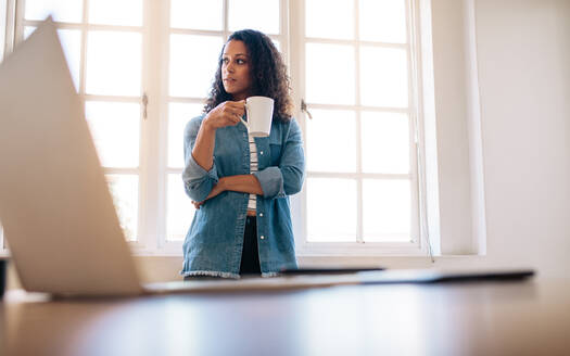 Eine Unternehmerin steht in einem Büro und hält eine Kaffeetasse, während sie wegschaut. Eine Frau steht in der Nähe ihres Tisches und trinkt Kaffee mit einem Laptop im Vordergrund. - JLPSF23337