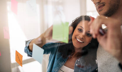 Ein Ehepaar schreibt auf Haftnotizen, die auf eine transparente Glaswand im Büro geklebt sind. Bürokollegen diskutieren Geschäftsideen und Pläne auf einer transparenten Glastafel. - JLPSF23339