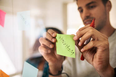 Man writing on sticky notes with marker pen and pasting on glass board in office. Entrepreneurs discussing business ideas and plans with sticky notes on glass wall. - JLPSF23340