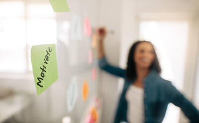 Unscharfes Bild einer Geschäftsfrau, die Haftnotizen an eine Glaswand klebt, mit Fokus auf ein darauf geschriebenes Post-it-Notizmotiv. - JLPSF23341