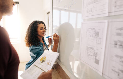 Geschäftsfrau schreibt auf Whiteboard im Büro, während ihr Partner Papiere hält. Bürokollegen diskutieren Geschäftsideen und Pläne auf einem Whiteboard im Büro. - JLPSF23342