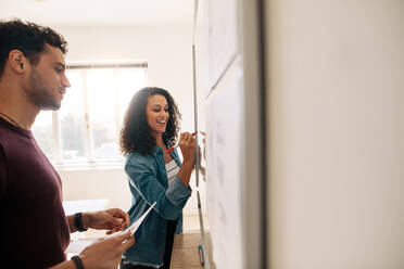 Smiling businesswoman writing on whiteboard in office while her partner is holding papers. Office colleagues discussing business ideas and plans on a whiteboard in office. - JLPSF23343