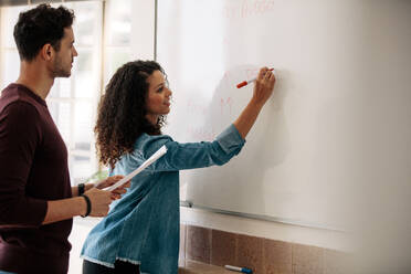 Office colleagues discussing monthly budget and plans on a whiteboard. Businesswoman writing on the board while her colleague holds a paper in hand. - JLPSF23352