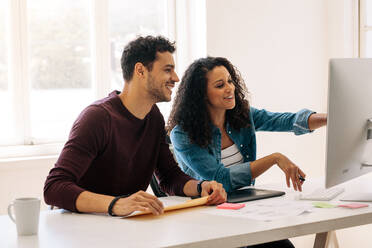 Businesswoman pointing towards the computer with a digital writing pad on the table. Woman entrepreneur discussing work with colleague sitting at her desk. - JLPSF23355