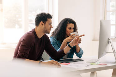 Businessman pointing towards the computer while discussing work with a business partner. Woman entrepreneur drinking coffee while discussing work with colleague sitting at her desk. - JLPSF23356