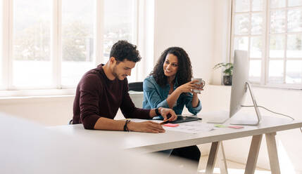 Businesswoman holding a coffee cup looking at her business partner while discussing work sitting at the table in office. - JLPSF23357