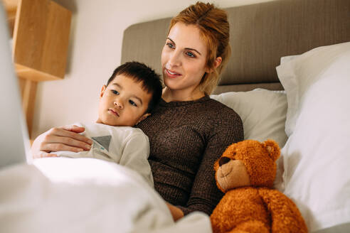 Schöne Mutter mit ihrem niedlichen Sohn auf dem Bett ruht etwas Interessantes auf dem Laptop beobachten. Unschuldiger Junge mit Mutter Blick auf Laptop mit einem Teddybär an der Seite. - JLPSF23368