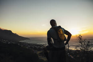 Rückansicht eines sportlichen Mannes, der auf einem Felsen sitzt und nach einem Trailrunning bei Sonnenuntergang eine Pause einlegt. Ein Mann entspannt sich auf einem Felsen neben einem Wanderweg auf einem Berg. - JLPSF23396