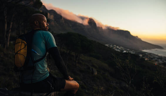 Rückansicht eines jungen Mannes, der auf einem Felsen sitzt und nach einem Trailrunning-Training eine Pause einlegt. Ein Mann sitzt auf einem Bergfelsen und schaut in den Sonnenuntergang. - JLPSF23398