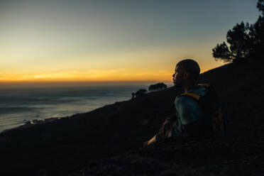 Sportler sitzt am Bergpfad und betrachtet den Sonnenuntergang. Mann ruht sich nach einem Trailrun aus und betrachtet die Landschaft. - JLPSF23399