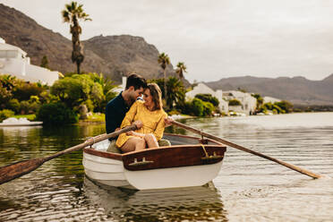 Young couple sitting together in a boat touching their heads. Couple having fun on a boat date in a lake with hills and houses in the background. - JLPSF23403