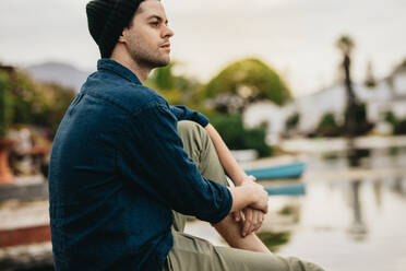 Side view of a man sitting near a lake looking away. Man relaxing on a holiday enjoying the scenic beauty of the lake. - JLPSF23406