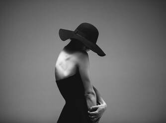 Artistic black and white portrait of sensual young woman standing against grey background. Elegant woman in black dress and hat. - JLPSF23409