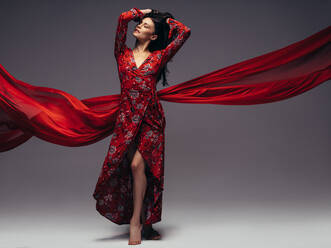Elegantes weibliches Modell in rotem Kleid mit einem fliegenden Tuch auf grauem Hintergrund. - JLPSF23444