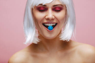 Nahaufnahme eines weiblichen Modells mit Make-up, das Minzbonbons auf rosa Hintergrund isst. Oben-ohne-Frau mit Perücke, mit einem Bonbon im Mund und geschlossenen Augen. - JLPSF23458