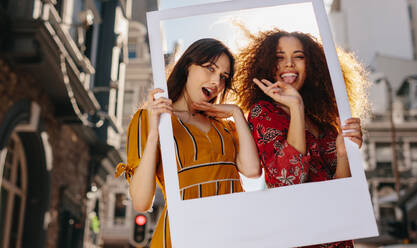 Two women wearing colourful dress standing outdoors holding a photo frame and making a funny facial expressions. Beautiful female friends enjoying outdoors with a blank picture frame. - JLPSF23474