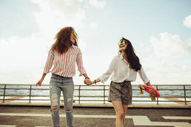 Freundinnen, die Hand in Hand an der Strandpromenade spazieren gehen. Zwei junge Frauen, die sich im Freien amüsieren. - JLPSF23488