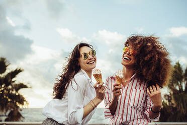 Zwei Freundinnen, die an einem Sommertag zusammen ein Eis essen. Fröhliche junge Frauen, die an der Strandpromenade ein Eis essen. - JLPSF23491