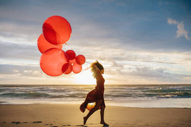 Young woman running along the beach holding a bunch of balloons. Female with balloons of different sizes at the beach sunset. - JLPSF23493