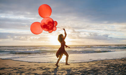 Totale Aufnahme einer jungen Frau, die mit Luftballons am Strand läuft. Eine Frau, die sich bei Sonnenuntergang am Meer amüsiert. - JLPSF23495