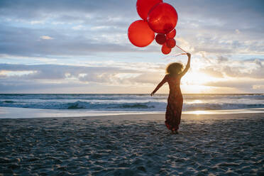 Rückansicht einer Frau, die am Strand spazieren geht und Luftballons in der Hand hält; eine Frau, die einen Tag am Meer genießt. - JLPSF23496