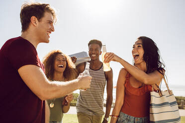 Eine Gruppe multiethnischer Freunde trinkt ein Bier, während sie zum Picknickplatz gehen. Männer und Frauen, die an einem Sommertag ein Picknick machen. - JLPSF23501