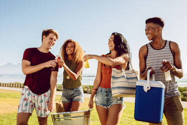 Group of people carrying cooler and beverage tub for party on beach. Diverse group of young people walking outdoors and having drinks. - JLPSF23504