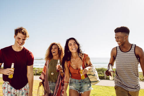 Eine Gruppe multiethnischer Freunde, die gemeinsam am Strand spazieren gehen, mit einem Bier in der Hand und lachend, eine sommerliche Strandparty. - JLPSF23506