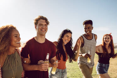 Multiracial group of young people having a great time together outdoor on a summer day. Young men and women with beers walking together. - JLPSF23517