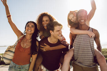 Two young men carrying female friends on their back and walking outdoors. Group of young friends having fun outdoors on a sunny day. - JLPSF23519